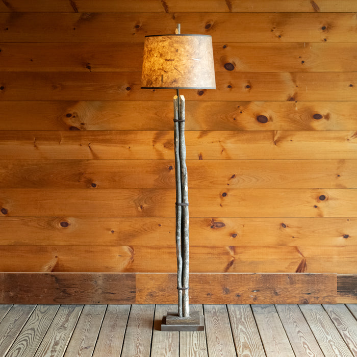 Front view of a rustic bundled wood floor lamp on a wide-plank wood floor against a rustic wooden wall 