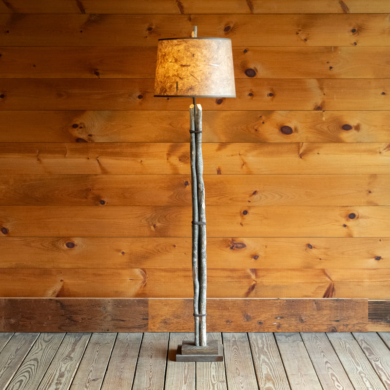 Front view of a rustic bundled wood floor lamp on a wide-plank wood floor against a rustic wooden wall 