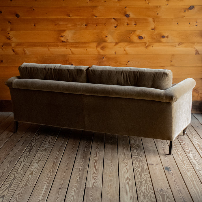 Angled back view of a rolled arm sofa upholstered in neutral mohair on a wide-plank wood floor against a rustic wooden wall