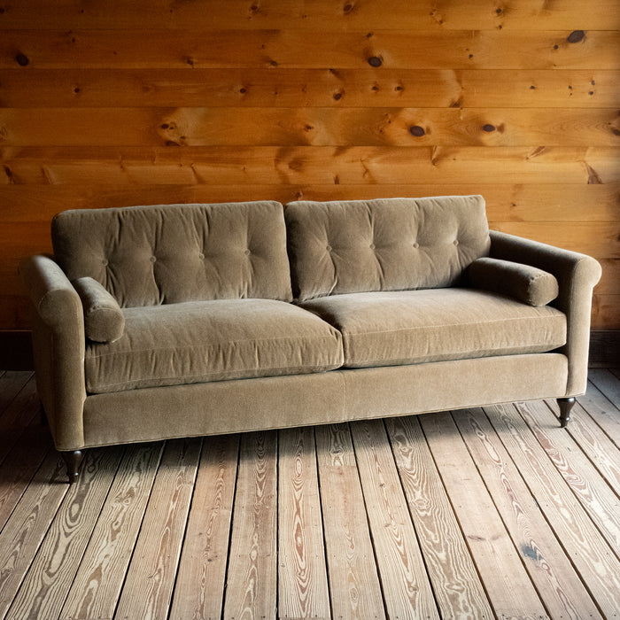 Angled front view of a rolled arm sofa upholstered in neutral mohair on a wide-plank wood floor against a rustic wooden wall
