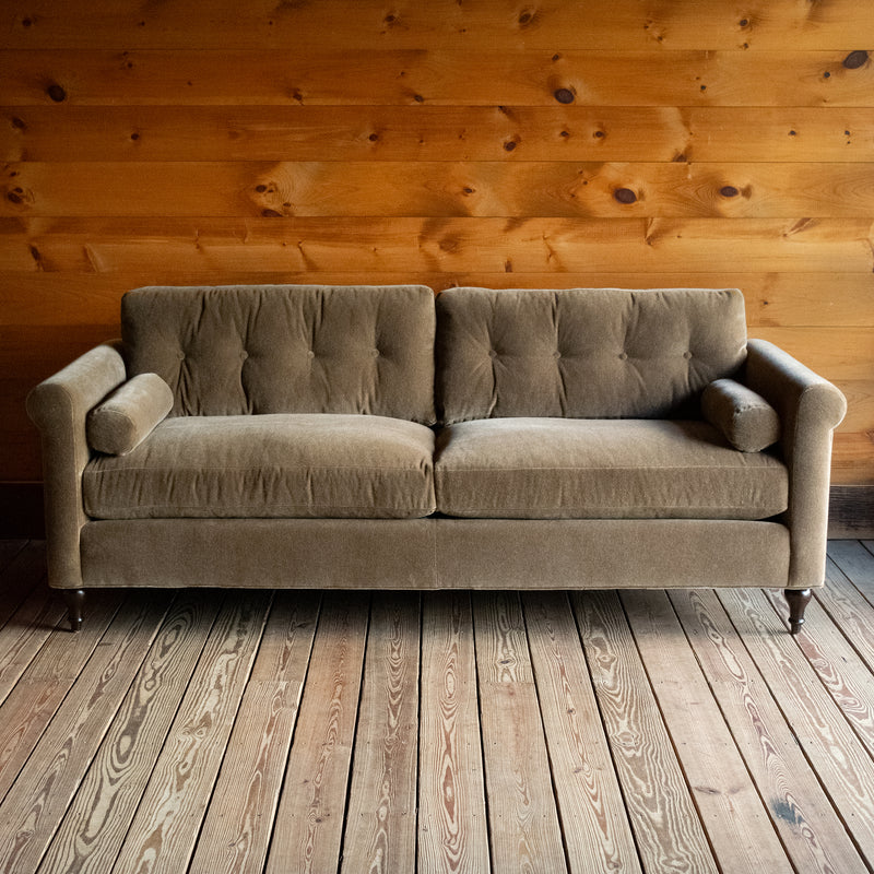 Front view of a rolled arm sofa upholstered in neutral mohair on a wide-plank wood floor against a rustic wooden wall