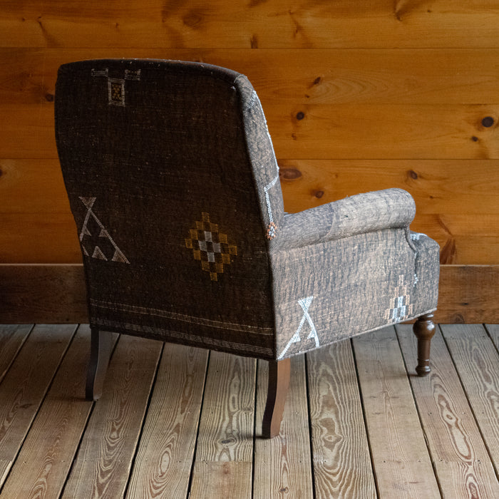 Angled back view of an armchair upholstered in dark brown Moroccan rug fabric featuring geometric motifs on a wide-plank wooden floor against a rustic wooden wall