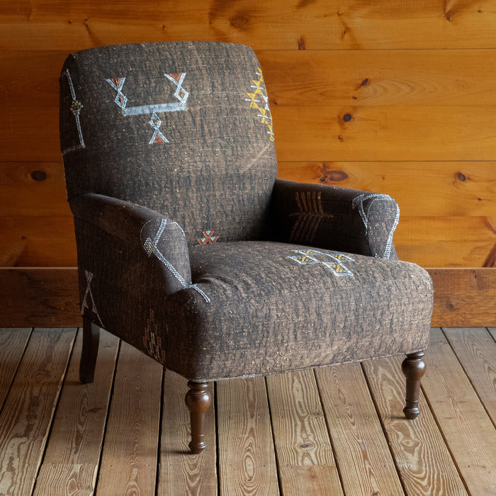Angled front view of an armchair upholstered in dark brown Moroccan rug fabric featuring geometric motifs on a wide-plank wooden floor against a rustic wooden wall