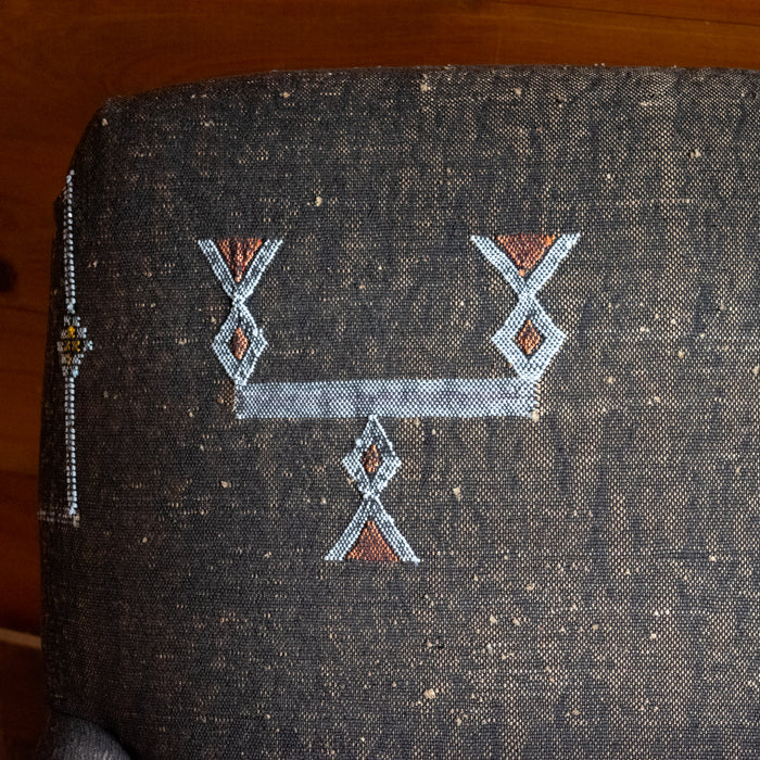 Close-up of the fabric of an armchair upholstered in dark brown Moroccan rug fabric with geometric motifs in white, yellow, and orange, against a rustic wooden wall