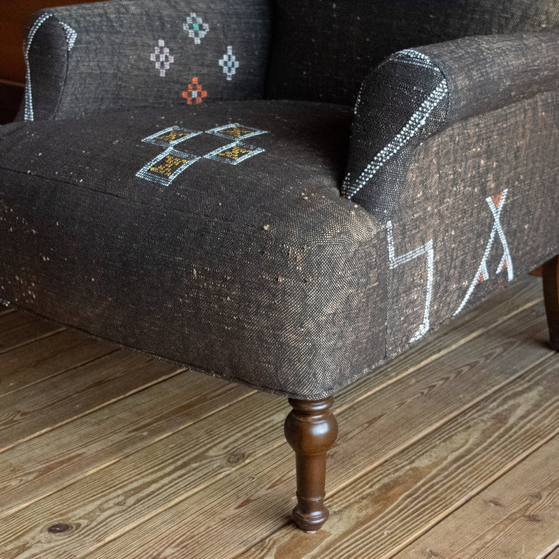 Close-up of the seat, arm, and leg of an armchair upholstered in dark brown Moroccan rug fabric with geometric motifs in white, blue, yellow, and orange, on a wide-plank wooden floor