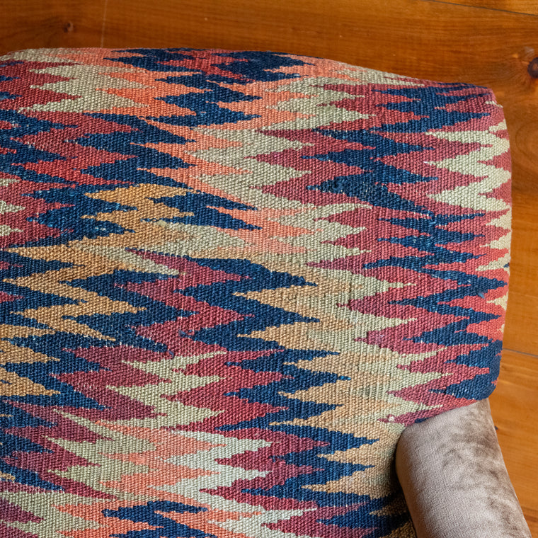 Angled back view of an armchair upholstered in a rug fabric featuring a rhythmic geometric pattern in warm shades of rust, red, and gold, set on a wide-plank wooden floor against a rustic wooden wall