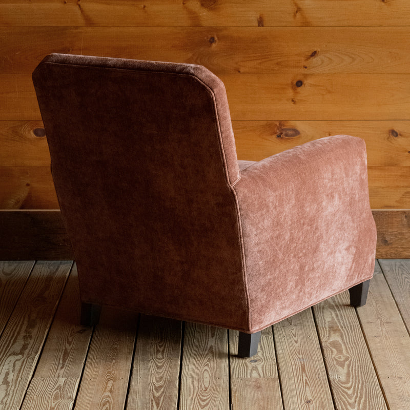 Angled back view of a cinnamon brown club chair with tapered hardwood legs and nailhead trim on a wide-plank wood floor against a rustic wooden wall 