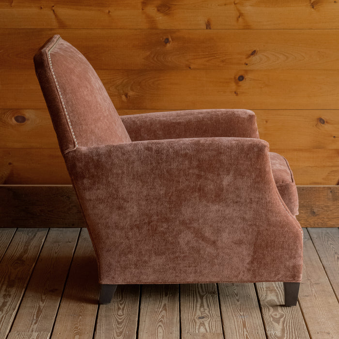 Profile view of a cinnamon brown club chair with tapered hardwood legs and nailhead trim on a wide-plank wood floor against a rustic wooden wall 