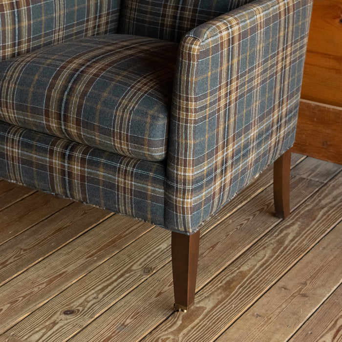 Close-up of the arm of a low, tight back chair with track arms upholstered in a rustic plaid fabric on a wide-plank wood floor against a rustic wooden wall