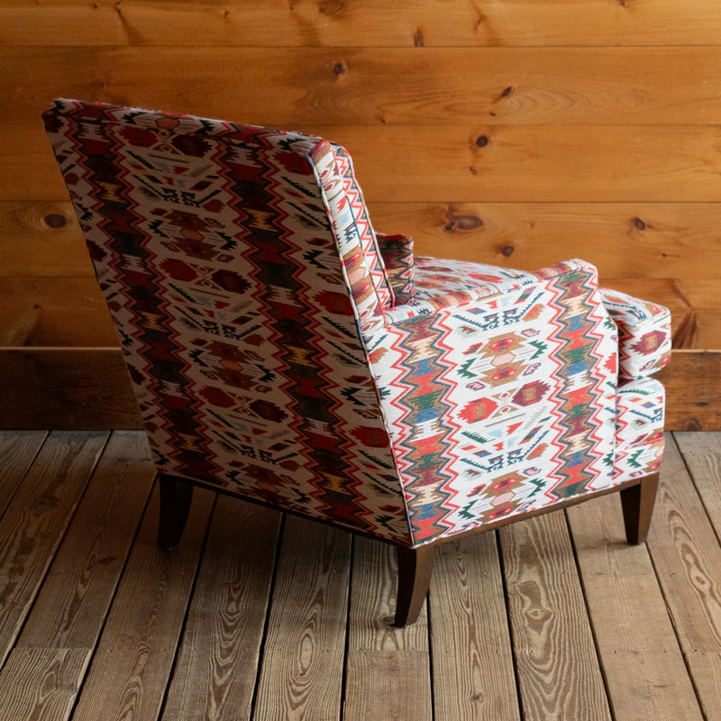 Angled back view of a rustic club chair upholstered in a vibrant rug-inspired fabric with a hardwood base and matching kidney pillow on a wide-plank wooden floor against a rustic wooden wall