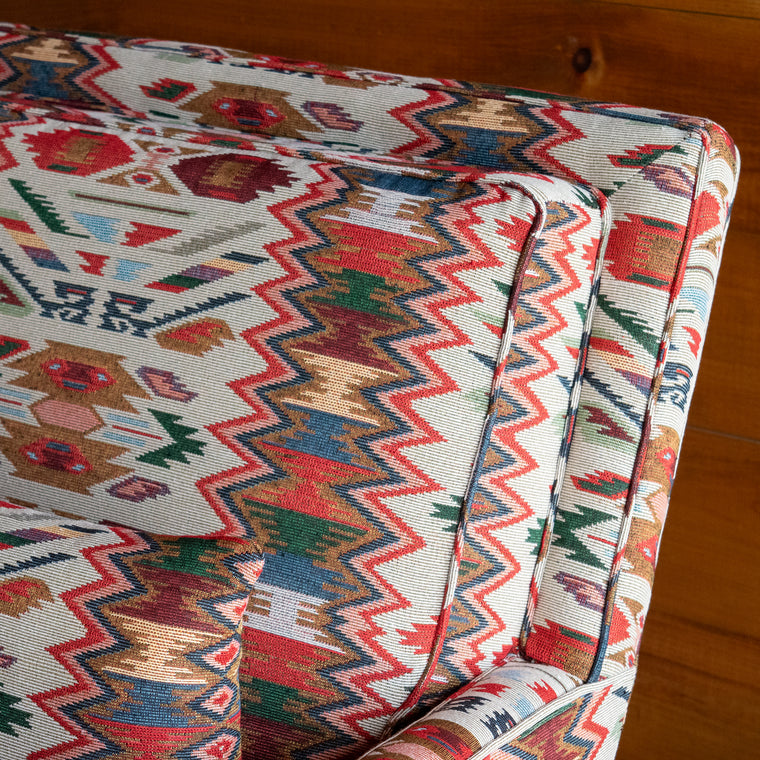 Angled back view of a rustic club chair upholstered in a vibrant rug-inspired fabric with a hardwood base and matching kidney pillow on a wide-plank wooden floor against a rustic wooden wall
