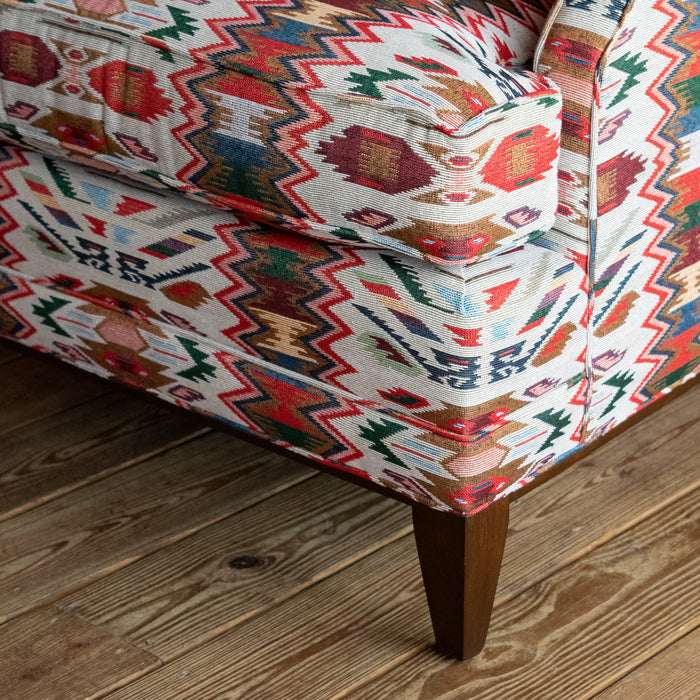 Close-up of the seat cushion and tapered hardwood leg of a rustic club chair upholstered in a vibrant rug-inspired fabric on a wide-plank wooden floor