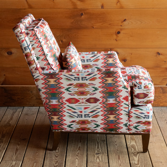 Profile view of a rustic club chair upholstered in a vibrant rug-inspired fabric with a hardwood base and matching kidney pillow on a wide-plank wooden floor against a rustic wooden wall
