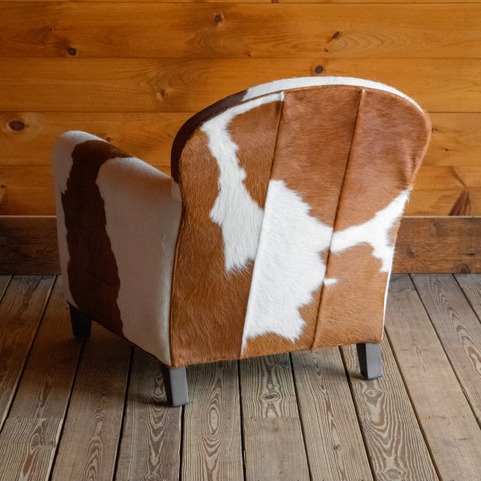 Angled back view of a brown and white cowhide armchair against a wooden wall