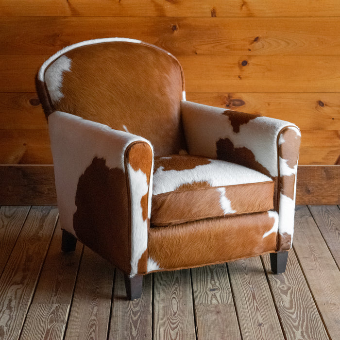 Angled front view of a brown and white cowhide armchair against a wooden wall