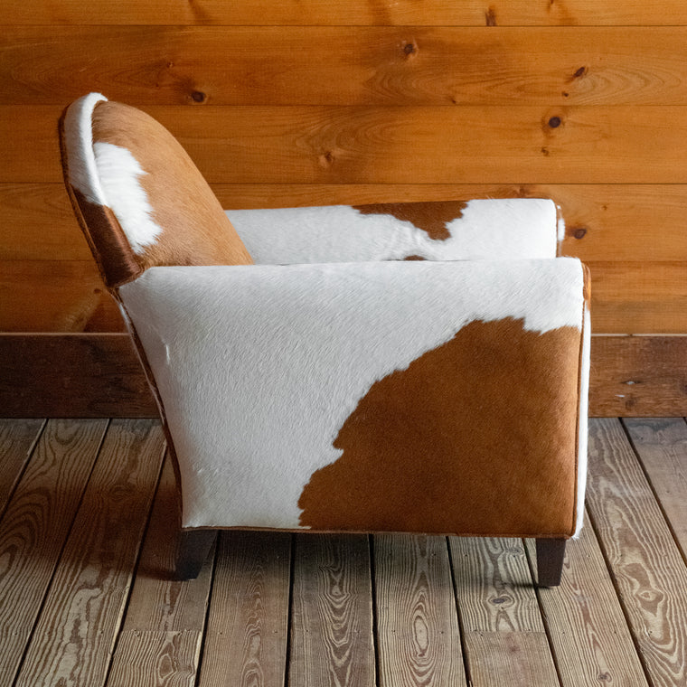 Angled back view of a brown and white cowhide armchair against a wooden wall