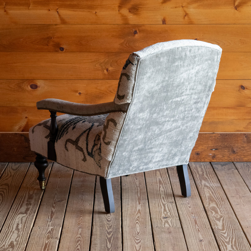 The back view of a lounge chair upholstered in a cream, blue, and sage green vintage rug-inspired fabric with turned front legs on casters against a wooden wall