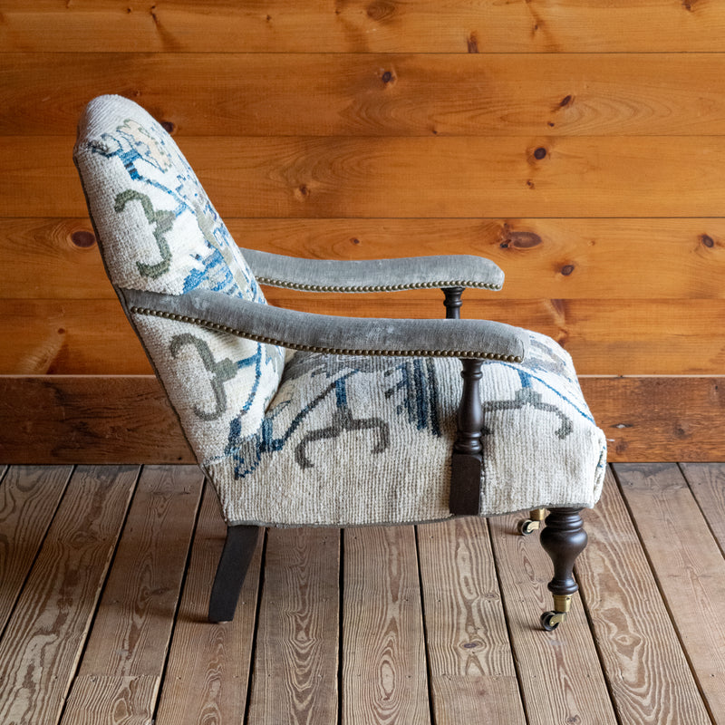 The profile view of a lounge chair upholstered in a cream, blue, and sage green vintage rug-inspired fabric with turned front legs on casters against a wooden wall