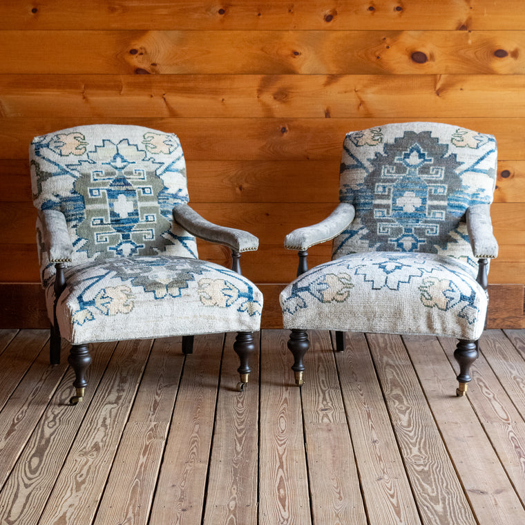The back view of a lounge chair upholstered in a cream, blue, and sage green vintage rug-inspired fabric with turned front legs on casters against a wooden wall