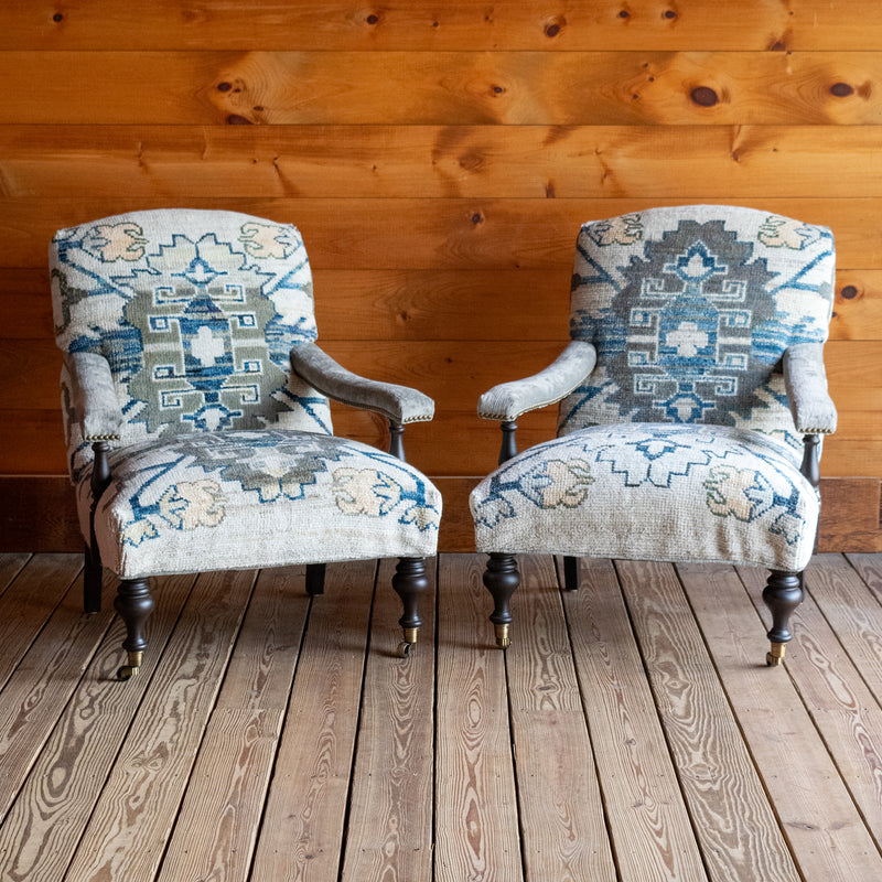 Two lounge chairs upholstered in a cream, blue, and sage green vintage rug-inspired fabric with turned front legs on casters against a wooden wall