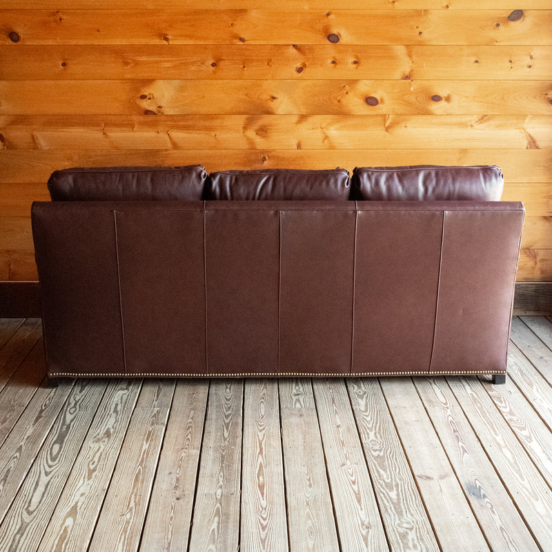 Back view of a dark brown leather three-seat sofa with nailhead trim against a wooden wall