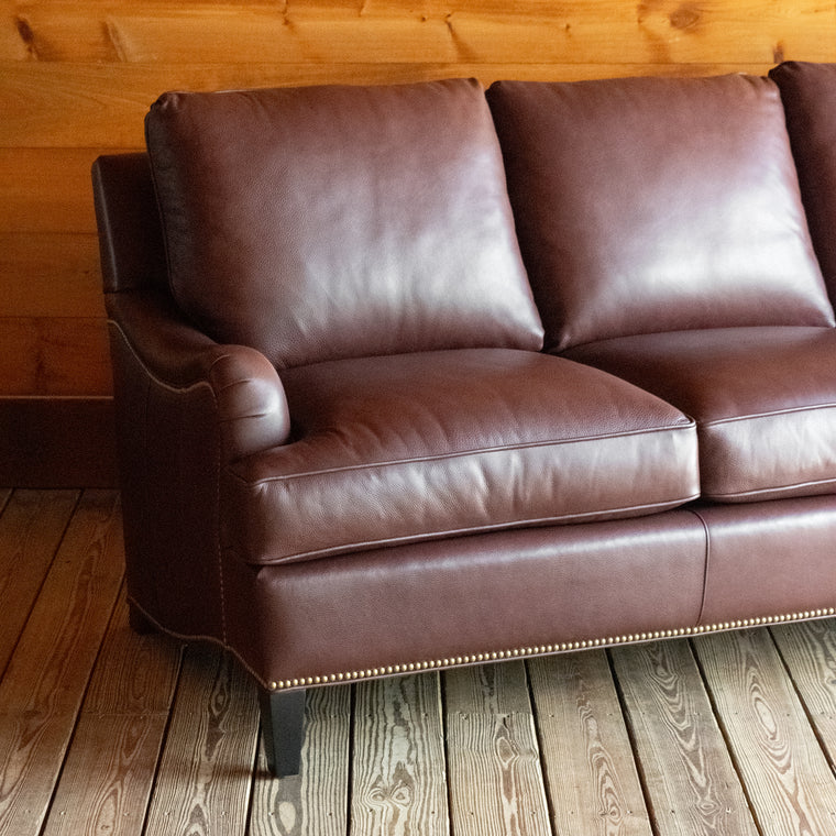Back view of a dark brown leather three-seat sofa with nailhead trim against a wooden wall