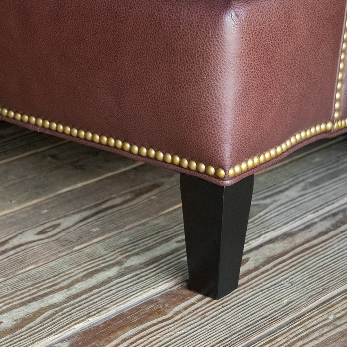 Leg close-up of a dark brown leather three-seat sofa with nailhead trim against a wooden wall