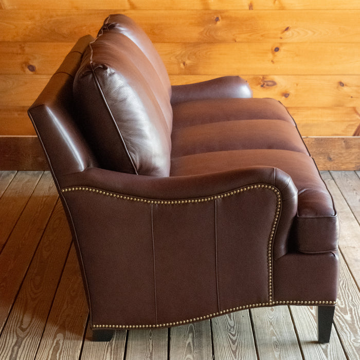 Pofile view of a dark brown leather three-seat sofa with nailhead trim against a wooden wall