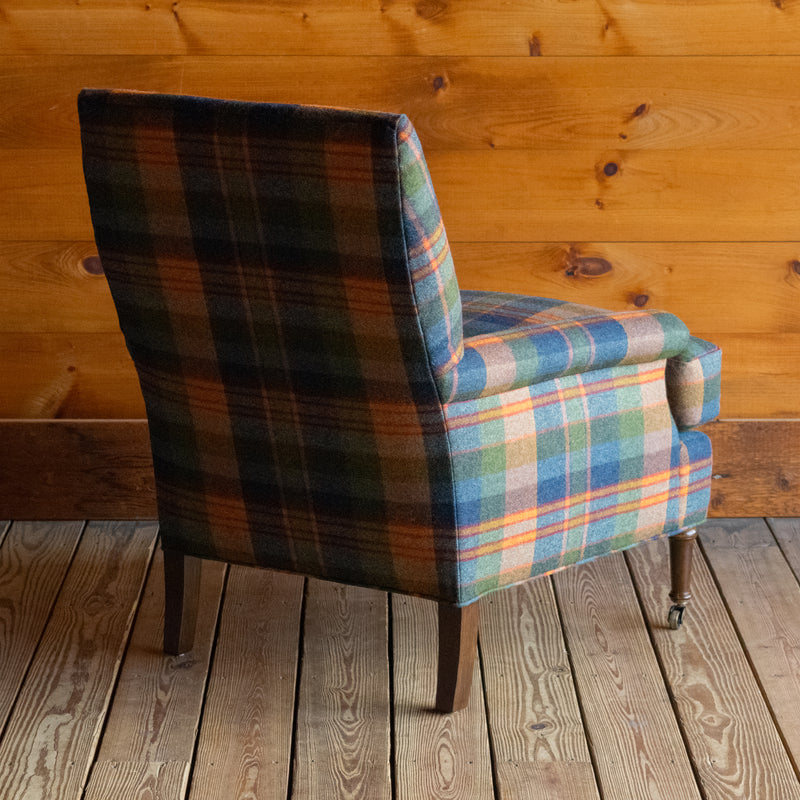 Angled back view of a tight back armchair in a vibrant wool plaid with slight rolled arms and turned legs on casters on a wide-plank wooden floor against a rustic wooden wall