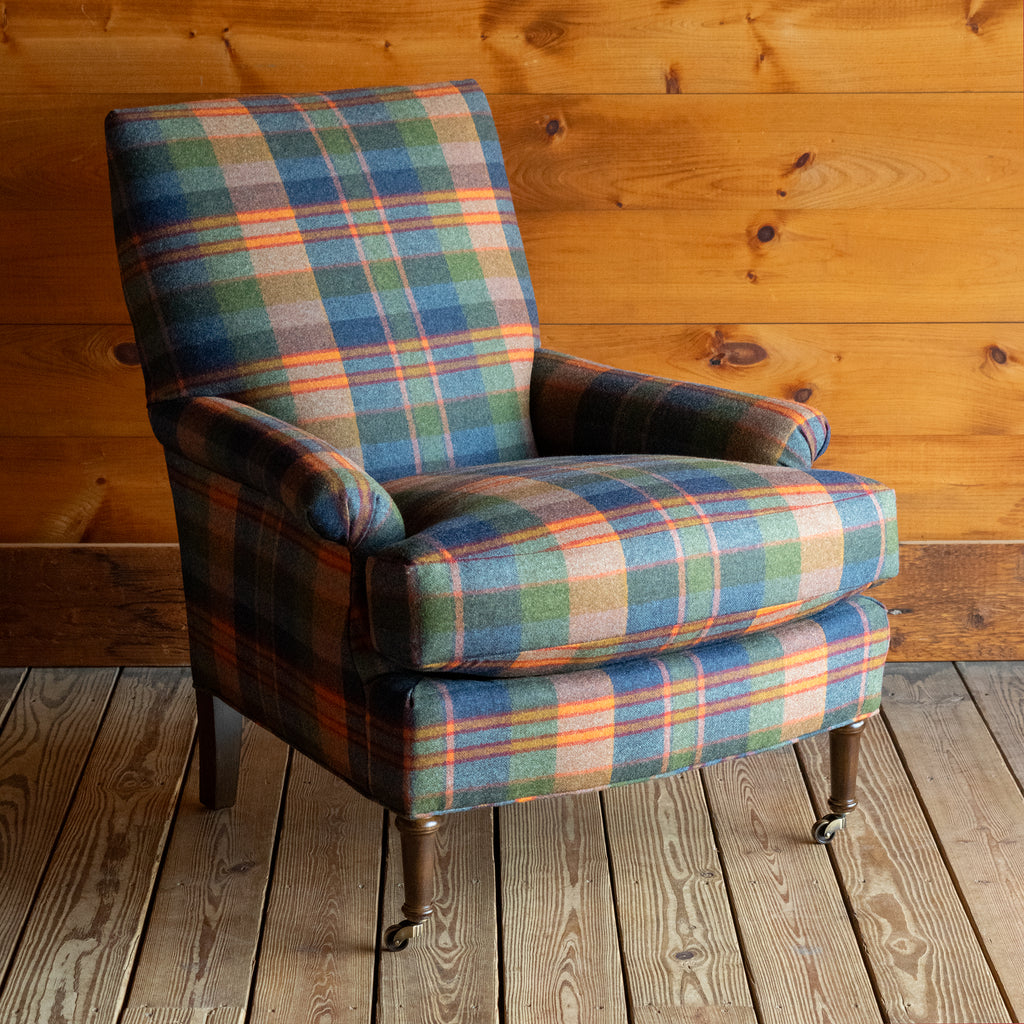 Angled front view of a tight back armchair in a vibrant wool plaid with slight rolled arms and turned legs on casters on a wide-plank wooden floor against a rustic wooden wall