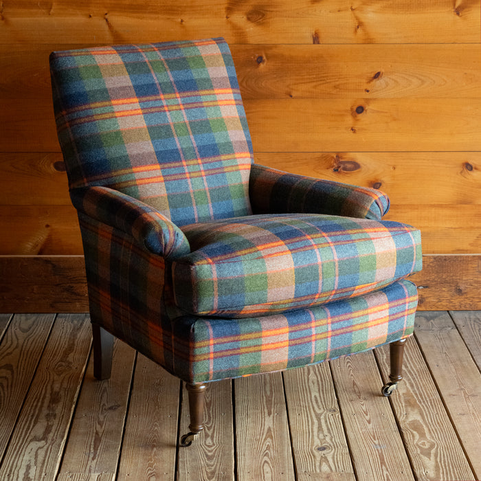 Angled front view of a tight back armchair in a vibrant wool plaid with slight rolled arms and turned legs on casters on a wide-plank wooden floor against a rustic wooden wall