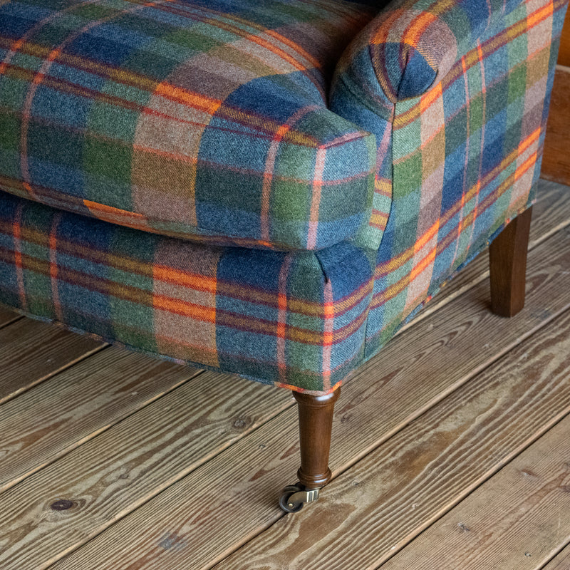Close-up of the slight rolled arm and turned leg of a tight back armchair in a vibrant wool plaid on a wide-plank wooden floor against a rustic wooden wall