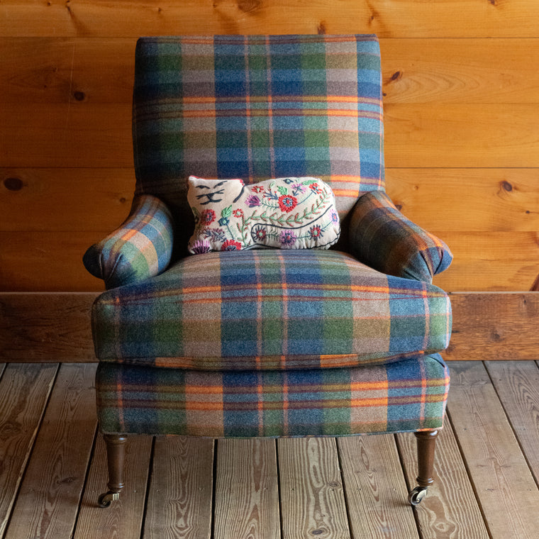 Angled back view of a tight back armchair in a vibrant wool plaid with slight rolled arms and turned legs on casters on a wide-plank wooden floor against a rustic wooden wall