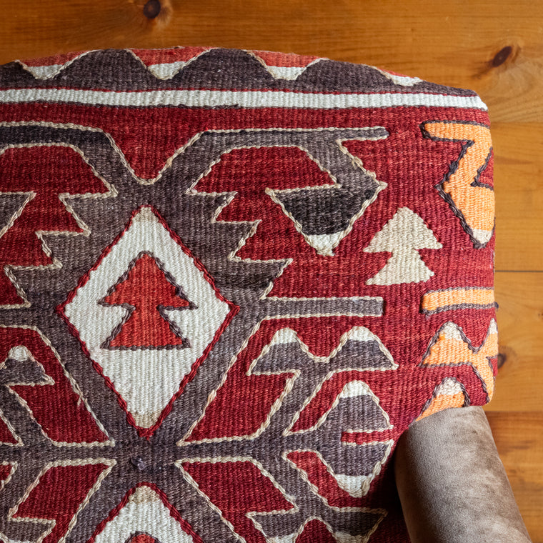 Angled back view of an armchair upholstered in a rug fabric featuring a large-scale geometric pattern on a saturated crimson background set on a wide-plank wooden floor against a rustic wooden wall