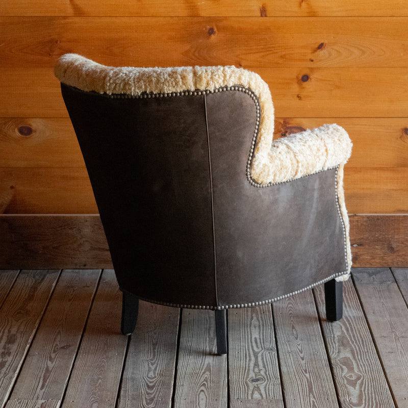 Angled back view of a golden shearling chair with contrasting brown suede leather back and sides against a wooden wall