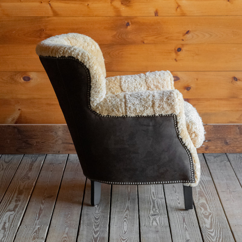 Profile view of a golden shearling chair with contrasting brown suede leather back and sides against a wooden wall