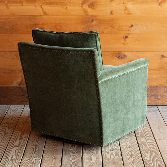 Angled back view of a swivel chair with track arms upholstered in a green velvet-chenille fabric on a wooden floor against a wooden wall
