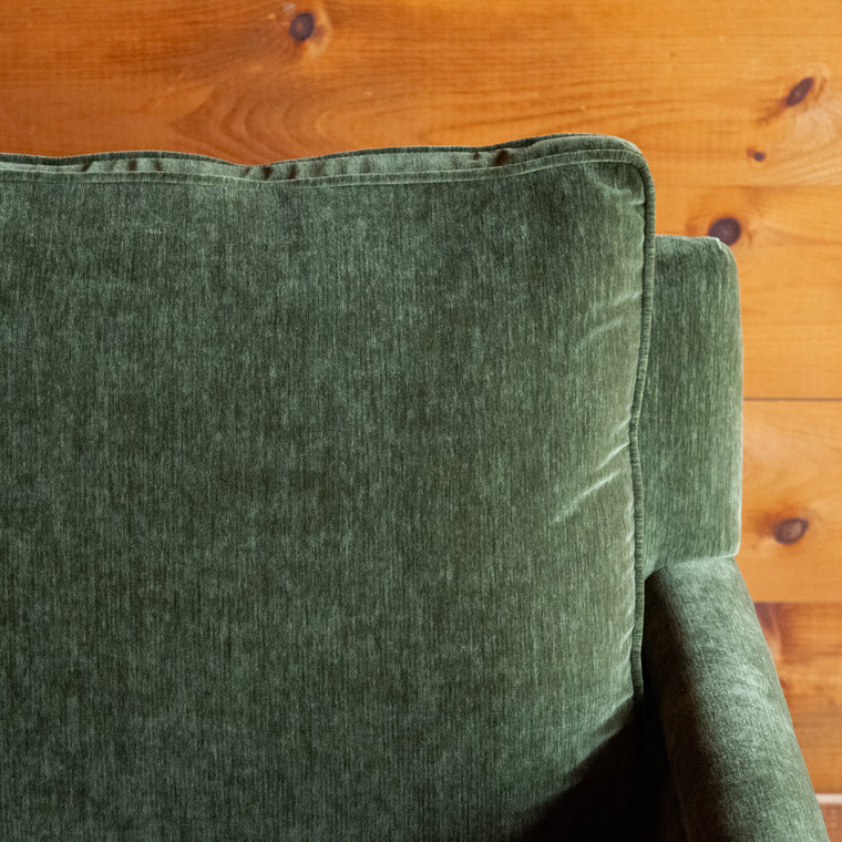 Angled back view of a swivel chair with track arms upholstered in a green velvet-chenille fabric on a wooden floor against a wooden wall