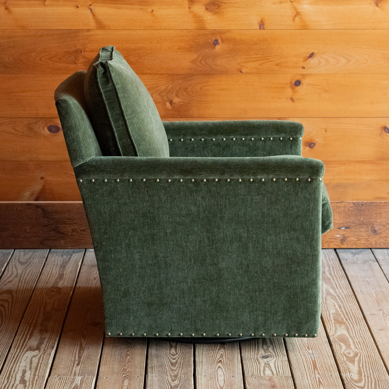 Profile view of a swivel chair with track arms upholstered in a green velvet-chenille fabric on a wooden floor against a wooden wall