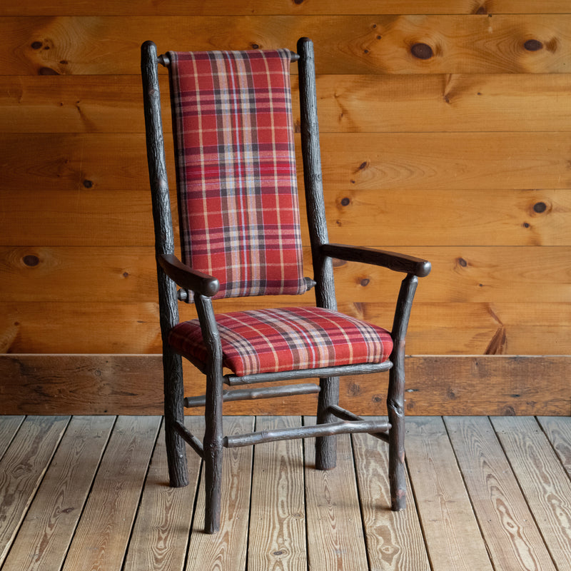 Angled front view of a hickory frame chair upholstered in a rustic red plaid fabric on a wide-plank wood floor against a rustic wooden wall