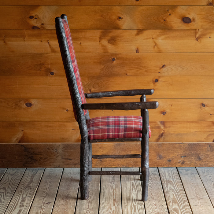 Profile view of a hickory frame chair upholstered in a rustic red plaid fabric on a wide-plank wood floor against a rustic wooden wall