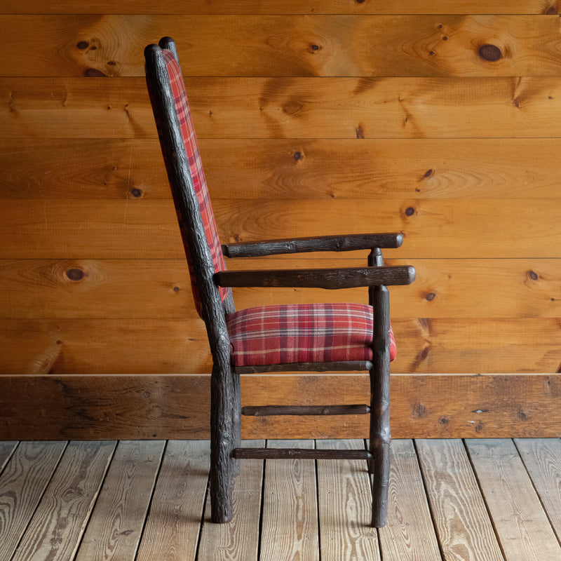 Profile view of a hickory frame chair upholstered in a rustic red plaid fabric on a wide-plank wood floor against a rustic wooden wall