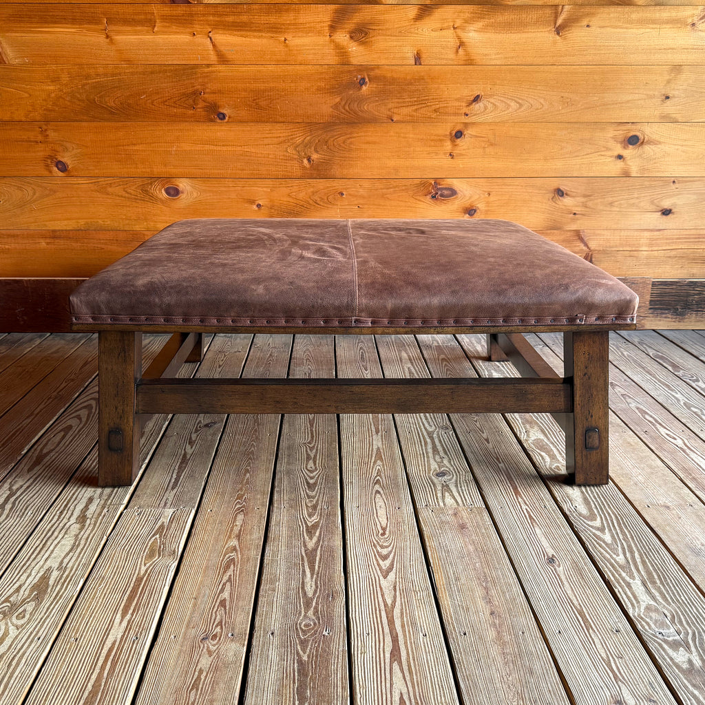Front view of a rustic leather ottoman with an open wooden frame against a wooden wall