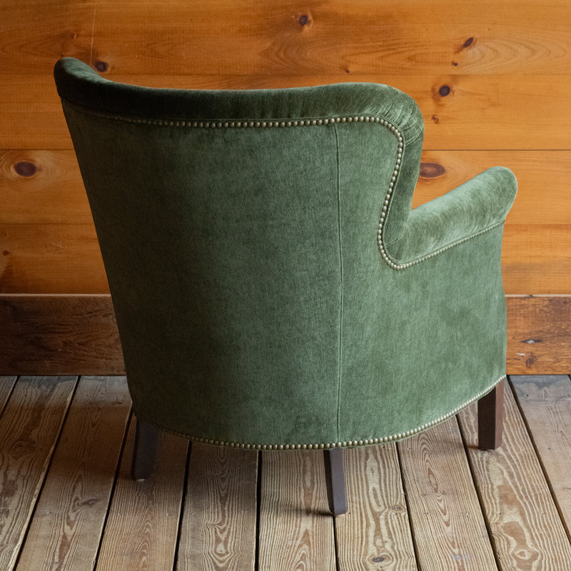 Angled back view of a large rustic arm chair upholstered in green velvet fabric on a wide-plank wooden floor against a rustic wooden wall