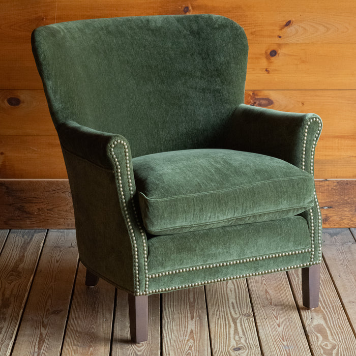 Angled front view of a large rustic arm chair upholstered in green velvet fabric on a wide-plank wooden floor against a rustic wooden wall