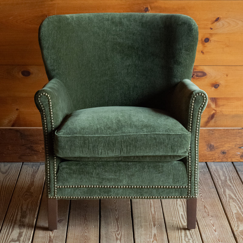 Front view of a large rustic arm chair upholstered in green velvet fabric on a wide-plank wooden floor against a rustic wooden wall