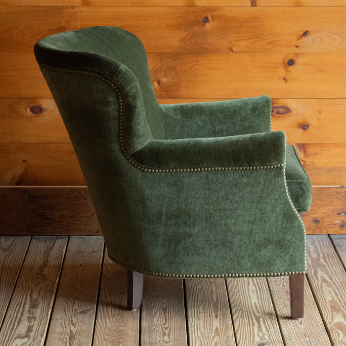 Profile view of a large rustic arm chair upholstered in green velvet fabric on a wide-plank wooden floor against a rustic wooden wall