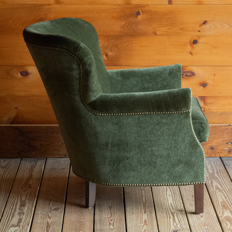 Profile view of a large rustic arm chair upholstered in green velvet fabric on a wide-plank wooden floor against a rustic wooden wall
