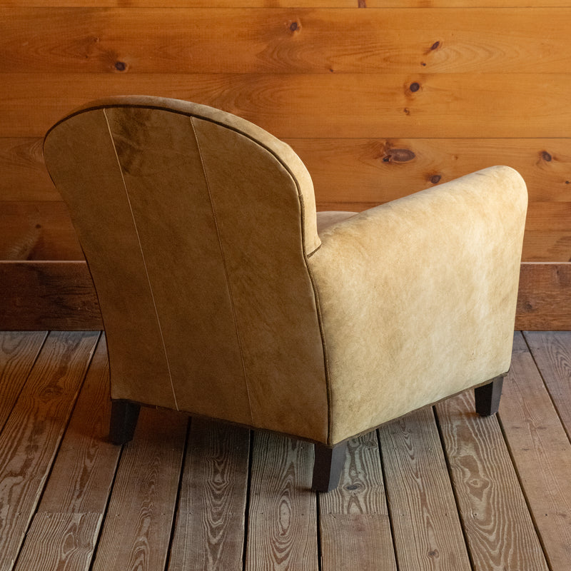Angled back view of a tight back leather arm chair upholstered in a rustic high-contrast leather with coordinating welt on a wide-plank wood floor against a rustic wooden wall