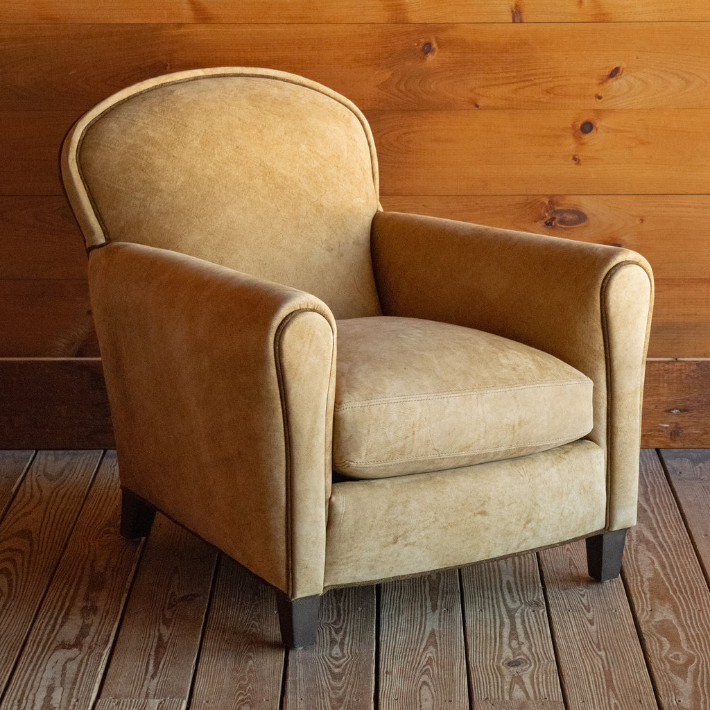 Angled front view of a tight back leather arm chair upholstered in a rustic high-contrast leather with coordinating welt on a wide-plank wood floor against a rustic wooden wall
